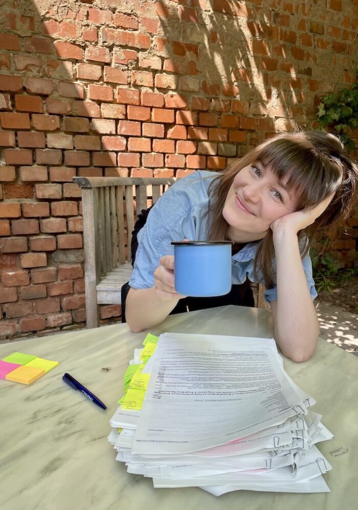 Braunhaarige Frau mit langen Haaren und Pony sitzt draußen, lächelt und hält eine Kaffeetasse hoch. Vor ihr auf einem Gartentisch liegt ein Stapel ausgedruckter wissenschaftlicher Artikel, die mit Post-It-Zetteln markiert sind. Im Hintergrund sind eine sonnenbeschienene Backstein-Mauer und Efeu zu sehen.