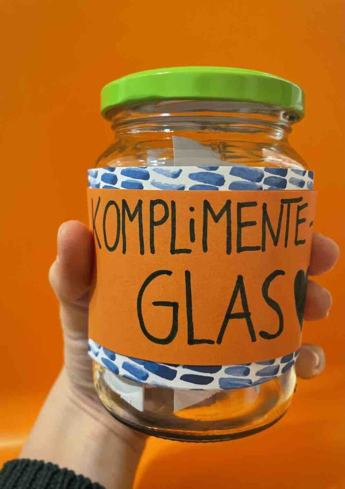 Foto eines Komplimenteglas: einfaches Schraubglas, beklebt mit hübschen bunten Zetteln mit der Aufschrift: "Komplimenteglas", darin gefaltete Zettel mit Komplimenten darin