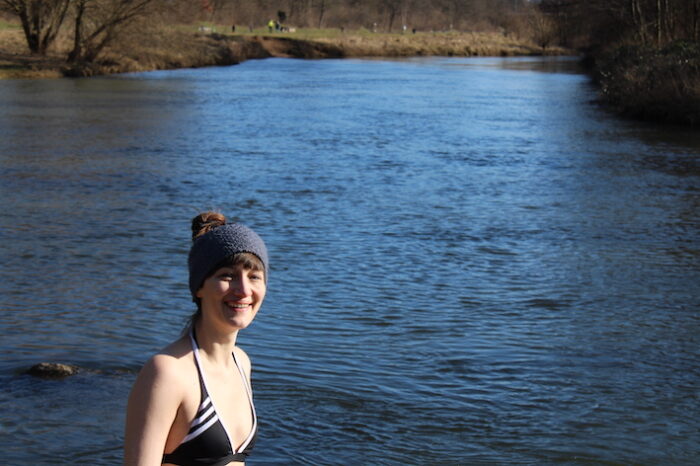 Frau beim Eisbaden. Sie trägt ein graues Stirnband und lächelt in die Kamera. Hinter ihr fließt ein tiefblauer Fluss. Es ist ein sonniger Wintertag.