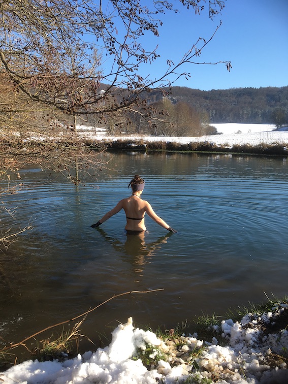 Frau beim Eisbaden. Sie steht hüfttief in einem Fluss und wartet darauf, dass sich ihr Körper an das kalte Wasser gewöhnt und sie losschwimmen kann. Die Landschaft drum herum ist verschneit.