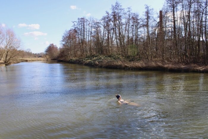 Eisbaden: Frau schwimmt im Winter in einem Fluss. Es ist ein sonniger Tag, sie hat sich auf den Rücken gedreht, ihr Gesicht wird von der Sonne angestrahlt. Im Hintergrund ist eine kahle Baumgruppe zu sehen.