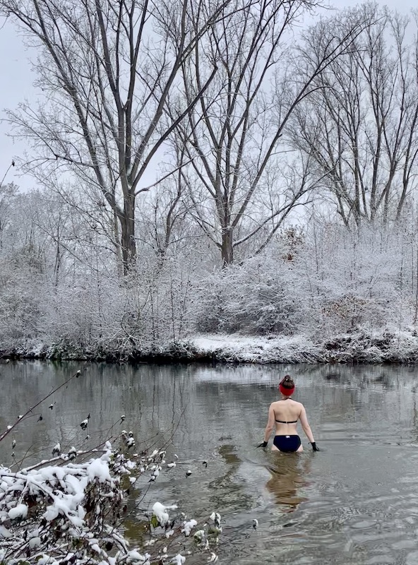 Eisbaden im Fluss: Zu sehen ist eine Frau im schwarzen Bikini und rotem Stirnband, die hüfttief in einem Fluss steht. Sie trägt schwarze Neoprenhandschuhe, um die Hände vor Kälte zu schützen. Um den Fluss herum stehen verschneite Bäume.